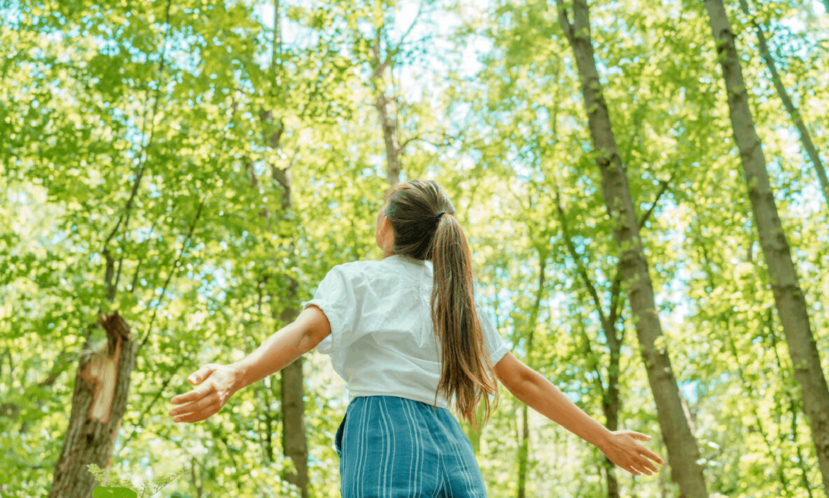 A woman standing in a sunlit forest with her arms outstretched, surrounded by tall green trees.